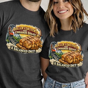 May include: Dark gray t-shirts with a graphic design featuring a roller coaster, cinnamon bread, and the text "ROLLER COASTERS AND CINNAMON BREAD A BALANCED DIET." The shirts are paired with blue jeans.