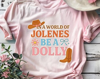 May include: A light pink t-shirt with the text "IN A WORLD OF JOLENES BE A DOLLY" in orange, blue, and pink. The shirt features a cowboy hat, a flower, a bow, and a cowboy boot graphic. A cowboy hat is on top of the shirt.