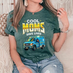 Op de afbeelding: Groen t-shirt met de tekst "COOL MOMS SAVE LIVES" in gouden en witte letters. Daaronder een blauwe ambulance-afbeelding. Het shirt is gemaakt van zacht materiaal en heeft korte mouwen.