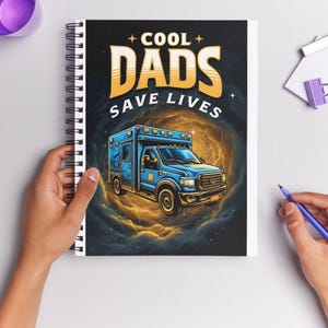 Op de afbeelding: Spiraalgebonden notitieboek met de tekst "COOL DADS SAVE LIVES" en een illustratie van een blauwe ambulance. De kaft heeft een donkere achtergrond met een wervelend gouden nevelontwerp.