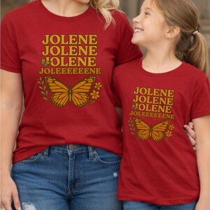 Op de afbeelding: Twee rode t-shirts met de naam "JOLENE" in gouden letters en een vlindergrafiek. De shirts worden gedragen met een blauwe jeans. De shirts hebben een vergelijkbaar ontwerp, wat wijst op een bijpassende set voor ouder en kind.
