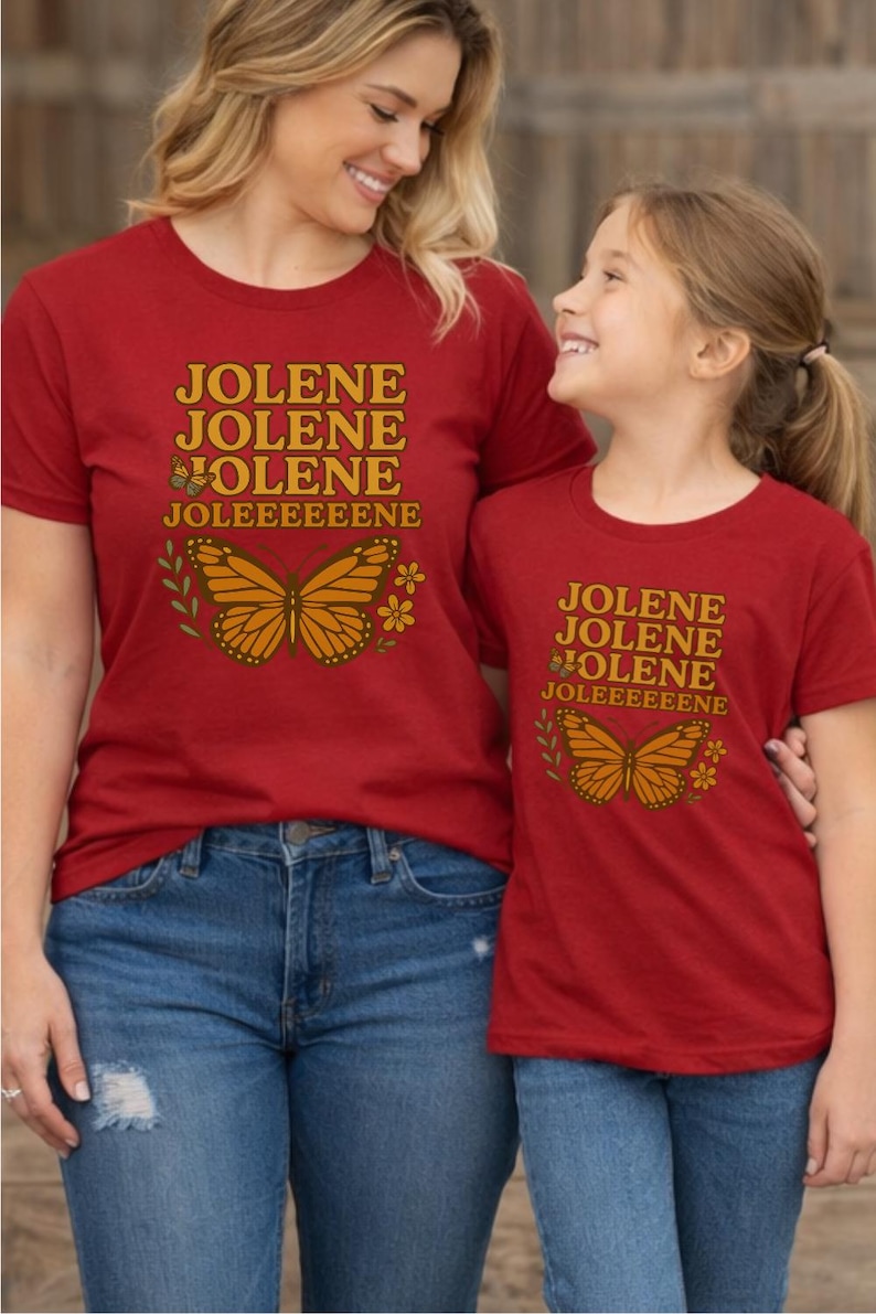 Op de afbeelding: Twee rode t-shirts met de naam "JOLENE" in gouden letters en een vlindergrafiek. De shirts worden gedragen met een blauwe jeans. De shirts hebben een vergelijkbaar ontwerp, wat wijst op een bijpassende set voor ouder en kind.