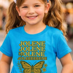 Op de afbeelding: Een turquoise t-shirt met de naam "JOLENE" in gouden letters, drie keer herhaald, en "JOLEEEEEEENE" onder een vlindergrafiek. Het t-shirt wordt gedragen door een jong meisje met een blauw-witte polkadot haarband.