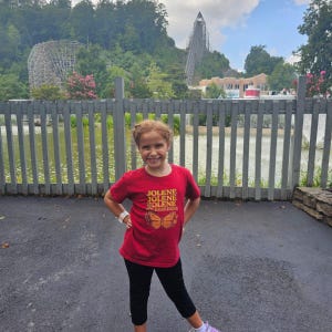 Op de afbeelding: Een jong meisje draagt een rood t-shirt met de tekst "JOLENE" herhaald en een vlinderontwerp. Ze staat voor een houten hek, met een achtbaan en bomen op de achtergrond.