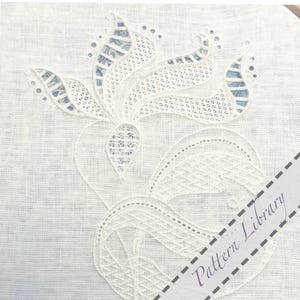 Puede incluir: Primer plano de un bordado de encaje blanco sobre una tela de color claro. El diseño presenta un motivo floral con detalles intrincados y patrones calados. La tela de fondo es de color claro y neutro, y el bordado es de estilo delicado y detallado.