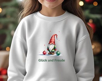 May include: A white sweatshirt featuring a watercolor illustration of a gnome with a red hat, green jacket, and a white beard. Two ornaments and the text "Glück und Freude" are also on the sweatshirt. The sweatshirt is worn by a young person.