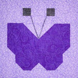 May include: Purple butterfly quilt block with a swirling pattern. The butterfly has two small black squares for antennae.