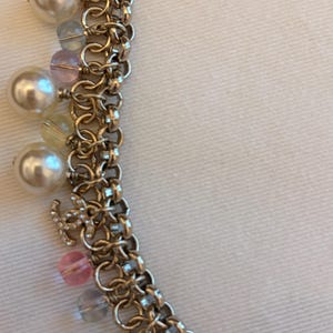 May include: A gold-tone chain necklace featuring a series of interlocking circular links. The necklace is adorned with dangling charms, including white faux pearls, translucent pink and blue beads, and a small gold-tone logo charm with crystals.