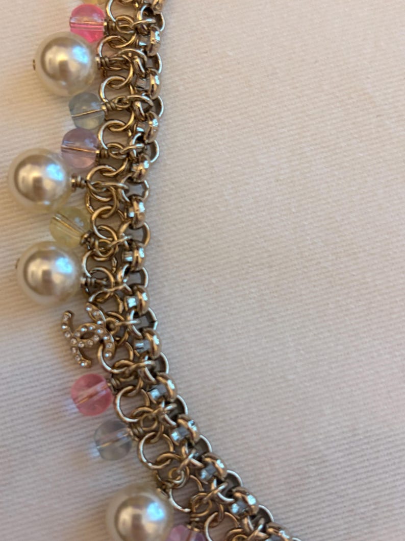 May include: A gold-tone chain necklace featuring a series of interlocking circular links. The necklace is adorned with dangling charms, including white faux pearls, translucent pink and blue beads, and a small gold-tone logo charm with crystals.