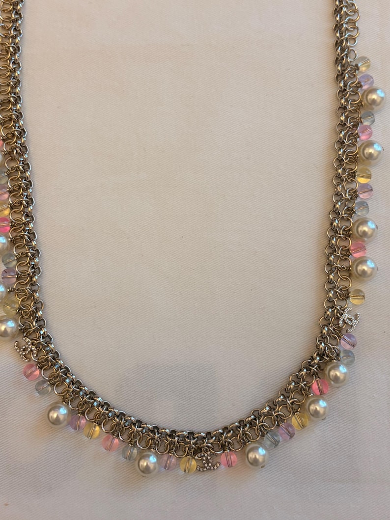 May include: A gold-tone chain necklace featuring multiple interlocking links adorned with dangling white faux pearls and translucent pink, yellow, and blue beads. Small silver-tone CC logo charms are interspersed throughout the chain design.
