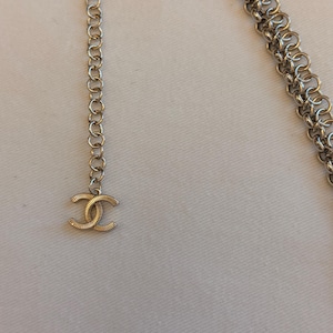 May include: A silver-toned metal chain belt featuring a series of interlocking circular links. The end of the chain terminates in a dangling, textured, double-C logo charm. The chain is constructed from multiple strands of small, uniform metal rings.