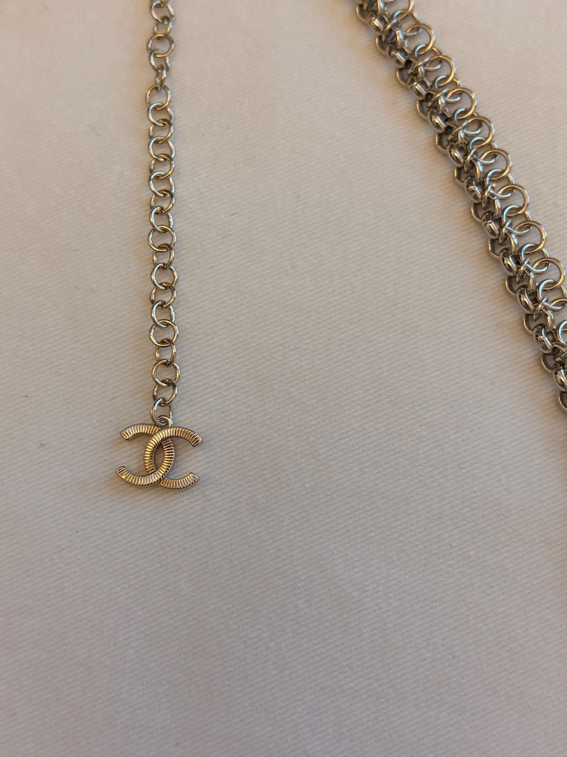 May include: A silver-toned metal chain belt featuring a series of interlocking circular links. The end of the chain terminates in a dangling, textured, double-C logo charm. The chain is constructed from multiple strands of small, uniform metal rings.