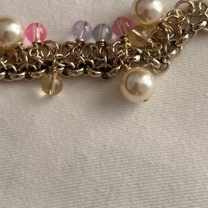 May include: A gold-toned chain bracelet featuring a series of interconnected metal rings. The bracelet is adorned with various dangling beads, including large cream-colored faux pearls, small translucent pink beads, light purple beads, and clear yellow beads.