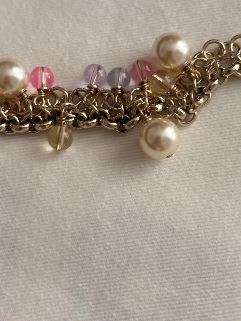 May include: A gold-toned chain bracelet featuring a series of interconnected metal rings. The bracelet is adorned with various dangling beads, including large cream-colored faux pearls, small translucent pink beads, light purple beads, and clear yellow beads.