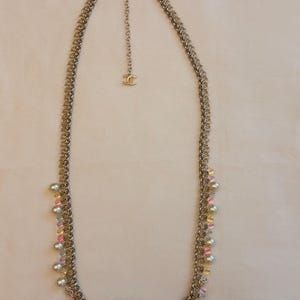May include: A long gold-tone chain necklace featuring a complex interlocking link design. The necklace is adorned with small white faux pearls and translucent pink and yellow beads hanging from the chain. A small interlocking CC logo charm is attached.
