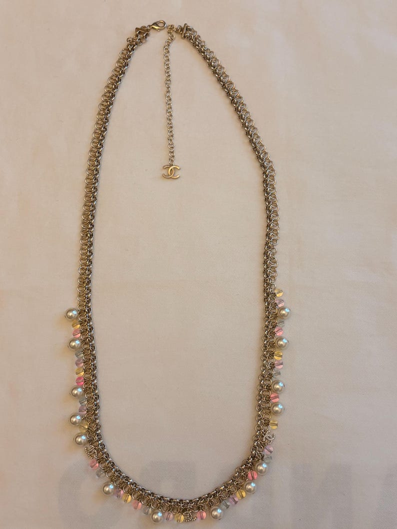 May include: A long gold-tone chain necklace featuring a complex interlocking link design. The necklace is adorned with small white faux pearls and translucent pink and yellow beads hanging from the chain. A small interlocking CC logo charm is attached.