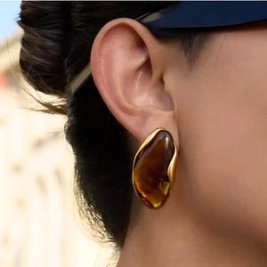 May include: A pair of gold-toned stud earrings with a unique, organic shape. Each earring features a large, amber-colored, irregular-shaped stone, set within a polished gold frame. The earrings have a smooth, glossy finish.