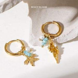 Pode incluir: Brincos de argola dourados com pingentes de concha e estrela do mar, adornados com contas azuis e pérolas. Os brincos são da Beach Bloom Jewelry, apresentados em fundo branco.