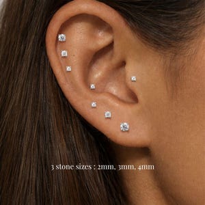 Può includere: Primo piano di un orecchio con diversi piccoli orecchini a bottone scintillanti. Gli orecchini sono di forma rotonda o quadrata e incastonati in un metallo di colore chiaro. Il testo in basso recita: "3 misure di pietra: 2mm, 3mm, 4mm."