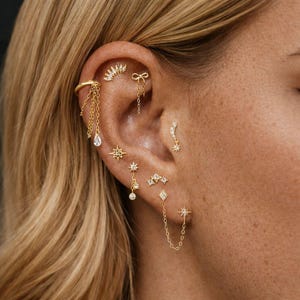 May include: An ear is decorated with a collection of gold earrings. The earrings feature designs such as a chain, a bow, a crescent moon, stars, and a diamond shape. Some earrings have clear, sparkling details. The ear is set against blonde hair.