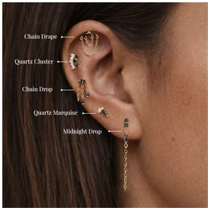 Può includere: Un orecchio adornato da molteplici orecchini dorati con dettagli in quarzo nero e catena. Gli orecchini includono modelli etichettati come "Chain Drap", "Quartz Cluster", "Chain Drop", "Quartz Marquise" e "Midnight Drop".