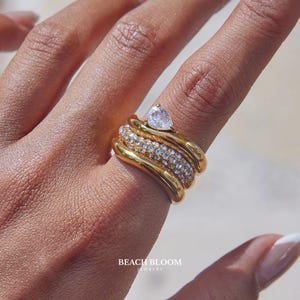 May include: A gold ring with a pear-shaped clear stone and multiple bands. The ring has a stacked design with a row of small, clear stones. The ring is on a finger. The text "BEACH BLOOM JEWELRY" is visible.