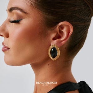 Puede incluir: Pendientes ovalados dorados con una piedra negra en el centro. Los pendientes tienen un borde texturizado y se muestran en una oreja. La imagen también incluye el texto "BEACH BLOOM JEWELRY".