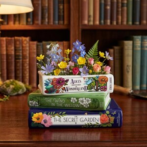 May include: A stack of three rectangular ceramic book-shaped planters. The top planter features the text Alice's Adventures in Wonderland, the middle reads The Wind in the Willows, and the bottom reads The Secret Garden, all filled with colorful flowers.