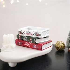 Pode incluir: Uma pilha de três recipientes de cerâmica em forma de livros retangulares. O livro branco superior diz Merry Christmas, o verde central diz Peace on Earth com um boneco de neve e o vermelho inferior diz Hope com detalhes florais.