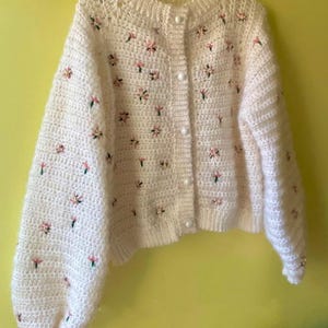 Puede incluir: Un cárdigan color crema tejido a mano con cuello redondo y mangas largas abullonadas. El suéter está adornado con pequeños detalles florales bordados en rosa y verde y tiene botones perlados.