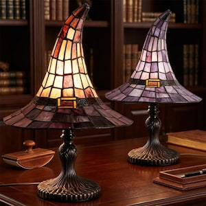 May include: Two stained glass table lamps shaped like pointed hats. The left lamp features warm orange and brown glass panels with a dark band and buckle. The right lamp displays purple and lavender glass panels with a similar dark band and buckle.