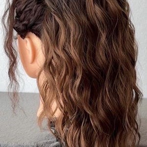May include: A wavy brown wig with dark roots and lighter brown highlights styled on a mannequin head. The hair features a side-swept section with visible texture and loose, flowing waves that cascade down the back and sides of the head.