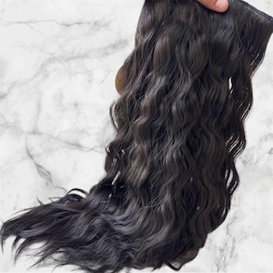 May include: Dark brown wavy hair extensions. The extensions are a deep, rich brown colour with a natural wave pattern. The hair appears thick and full, with a slight sheen. The extensions are held up by a hand, showing the attachment method.