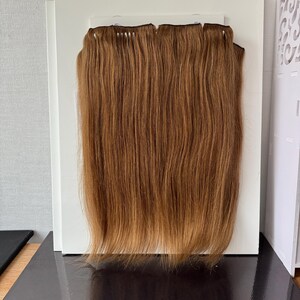 Op de afbeelding: Een set steile hairextensions in een medium goudbruine tint. Het haar is bevestigd aan een horizontale baan aan de bovenkant, met fijne, gladde lokken die verticaal hangen in een uniforme, strakke en lange stijl met natuurlijke highlights.