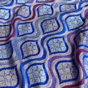 May include: A fabric featuring a repeating geometric pattern of wavy lines in shades of blue and purple. Each enclosed section contains a detailed, light-colored floral motif. The textile has a textured appearance with a consistent, structured design.