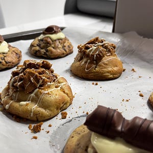 May include: Several round baked cookies arranged on white parchment paper. Some cookies are topped with white icing and a chocolate bar piece, while others feature a golden-brown crumbled topping drizzled with white icing and scattered crumbs.