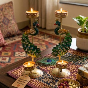 May include: Two gold-toned metal candle holders featuring a peacock design with green and blue enameled tail feathers. Each holder has a circular base and a top cup for a tea light candle. A paper tag attached to one base reads Handcrafted Meenakari Peacocks.