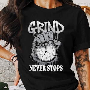 May include: Black t-shirt with the words "GRIND NEVER STOPS" in white. A hand is holding a stopwatch with the time displayed. The shirt is a crew neck and has short sleeves.