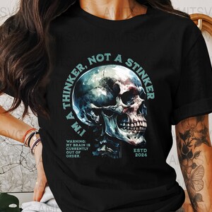 Może przedstawiać: Czarny t-shirt z szczegółową grafiką czaszki i napisem "I'M A THINKER, NOT A STINKER." Projekt zawiera rok "ESTD 2024" i ostrzeżenie "MY BRAIN IS CURRENTLY OUT OF ORDER."