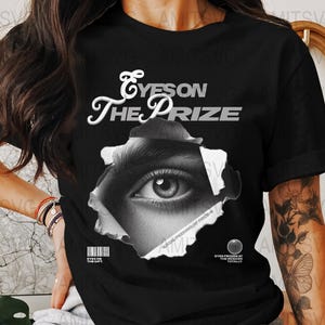 Pode incluir: Camiseta preta com o texto "Eyes on the Prize" em escrita branca. A camiseta apresenta uma imagem em tons de cinza de um olho espreitando por um efeito de papel rasgado. Um código de barras e texto adicional são visíveis.