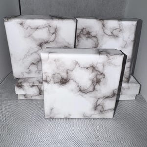 Puede incluir: Varias cajas blancas con un patrón de mármol. Las cajas son rectangulares y de diferentes tamaños, con una mezcla de cajas grandes y pequeñas. El diseño de mármol presenta vetas grises y marrones sobre un fondo blanco.