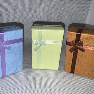 Pode incluir: Três caixas de presente retangulares em tons de azul, verde e castanho. Cada caixa é adornada com uma fita e um laço em cores complementares. As caixas estão dispostas lado a lado, sugerindo uma coleção de presentes.