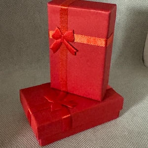 Pode incluir: Duas caixas de presente vermelhas, uma retangular e outra quadrada, estão empilhadas. Ambas as caixas são adornadas com fitas e laços vermelhos. As caixas são provavelmente para joias ou pequenos presentes.