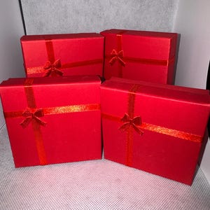 Pode incluir: Quatro caixas de presente vermelhas com fitas e laços vermelhos a condizer. As caixas são quadradas e têm um acabamento brilhante. Os laços estão centrados na parte superior de cada caixa.