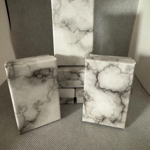 Pode incluir: Várias caixas de presente retangulares com um padrão de mármore branco. As caixas estão dispostas em grupo, algumas em pé e outras planas. O design de mármore apresenta veios cinzentos.