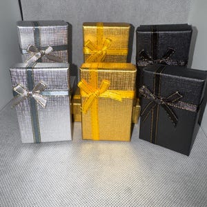 Pode incluir: Seis pequenas caixas de presente em prata, ouro e preto. Cada caixa é quadrada com um padrão texturizado e um laço de fita a condizer. As caixas prateadas têm detalhes azuis, as douradas laços amarelos e as pretas laços pretos.