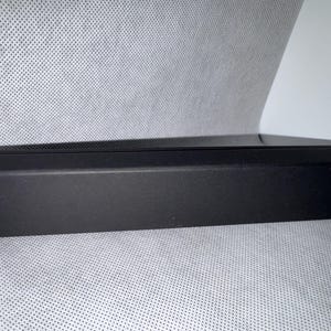 Pode incluir: Uma caixa retangular preta com uma parte superior texturizada e laterais anguladas. A caixa é feita de um material fosco e parece estar vazia. A caixa está em uma superfície cinza claro.