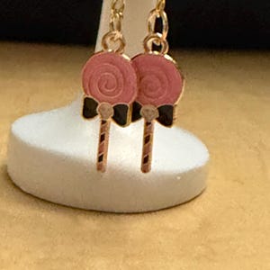 Peut inclure: Une paire de boucles d'oreilles pendantes dorées représentant une sucette rose avec un motif en spirale, un petit nœud noir à la base et un bâton rayé. Les boucles d'oreilles sont suspendues à des crochets dorés pour oreilles percées.