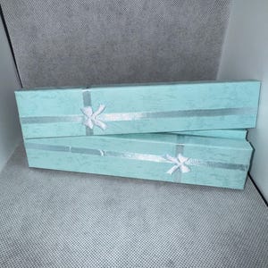 Pode incluir: Duas caixas de presente retangulares azul claro com fita prateada e laços brancos. As caixas estão empilhadas uma sobre a outra, sobre um fundo texturizado cinza claro.