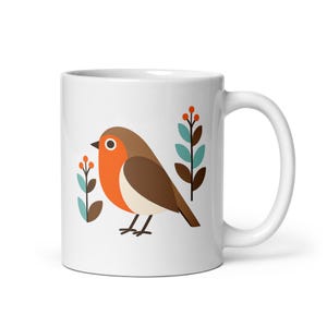Könnte beinhalten: Ein weißer Keramikbecher mit einer grafischen Illustration eines Rotkehlchens mit braunem Rücken, orangefarbener Brust und cremefarbenem Bauch. Der Vogel wird von zwei stilisierten botanischen Stängeln mit blaugrünen Blättern und Beeren flankiert.